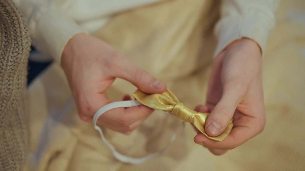 Close up of hands holding and adjusting small golden bow tie with elastic band. person wears light colored clothing focusing on delicate manipulation of fabric texture and form against soft