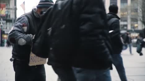 a homeless man stands on a city street preparing his sign as people pass by. the hustle of urban life is evident as pedestrians walk quickly, absorbed in their own activities.
