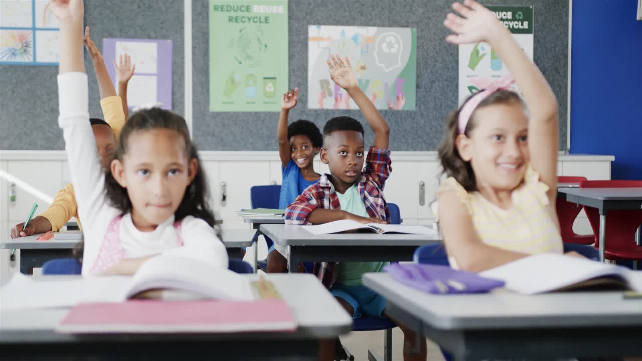 Happy diverse schoolchildren at desks raising hands in classroom at elementary school