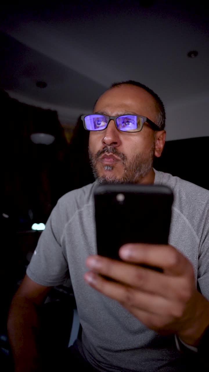 South asian man using smartphone in dark room wearing blue light filter glasses for reducing eye strain and improving sleep