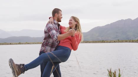 caucasian couple having a good time on a trip to the mountains, dancing near lake and smiling