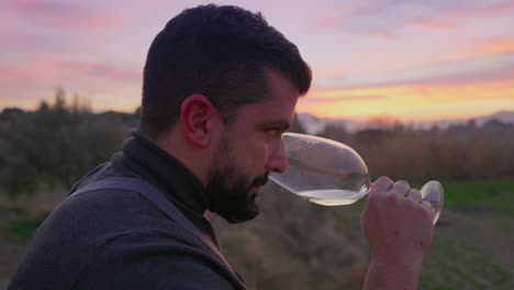 Un-Barbudo-Huele-Y-Prueba-Una-Copa-De-Vino-Blanco-Al-Atardecer-Bajo-Un-Cielo-Primaveral-De-Naranjas-Y-Rosas,-Con-Un-Prado-Verde