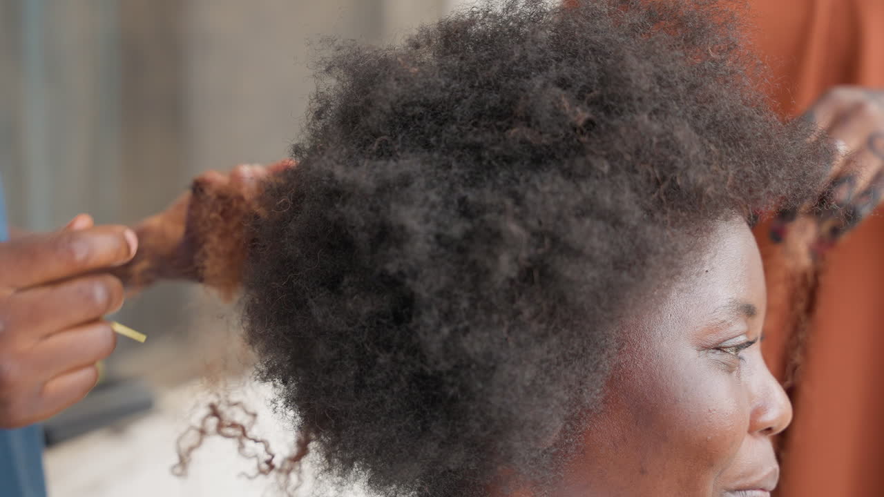 Donna di profilo con capelli afro, stilista che districa e separa delicatamente i riccioli usando pettine e dita, rivelando texture e movimento, ambiente interno caldo, tecnica paziente che trasforma sezioni aggrovigliate