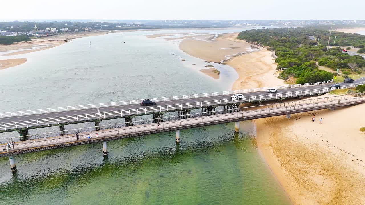 Drone footage captures vehicles crossing Barwon Heads Bridge over a serene river, with sandy shores and lush greenery in view