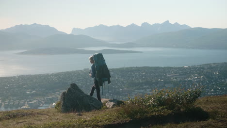 beautiful hiker girl with heavy backpack, standing on a cliff and enjoying the spectacular view of tromsø, sunny summer day in norway
