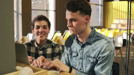 two young people are smiling, listening to music in their headphones and pointing at something on laptop screen. attractive female, handsome male in their 20's, leisure time.