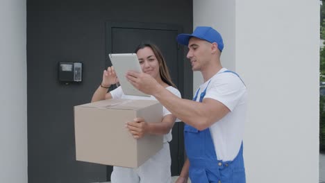 beautiful woman meets a delivery man who gives her a parcel box beside her home, she signs electronic signature on tablet. concept of courier, home delivery, online shopping