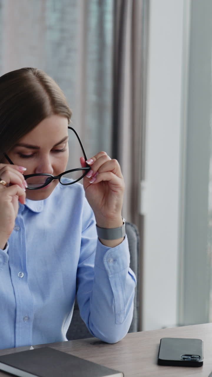 Long-haired woman in blue shirt sitting at desk in office. Smiling lady puts on glasses. Close up. Vertical video