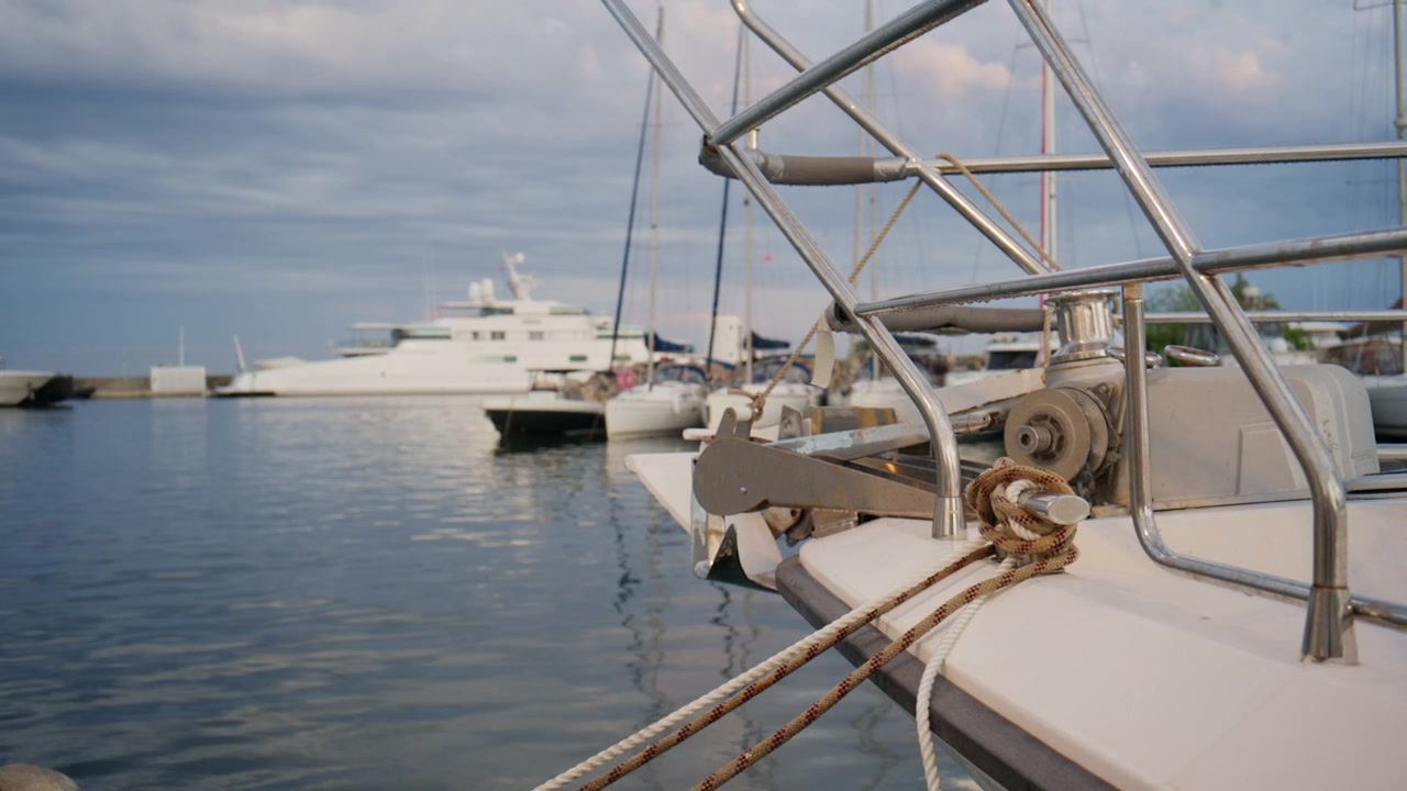 Primo piano della prua di una barca a vela, ancora, cime e ringhiere metalliche al porto turistico di Port Vauban ad Antibes, con yacht e acqua calma del Mediterraneo sullo sfondo.