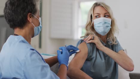 Senior-african-american-female-doctor-vaccinating-female-patient-at-home-both-wearing-face-masks