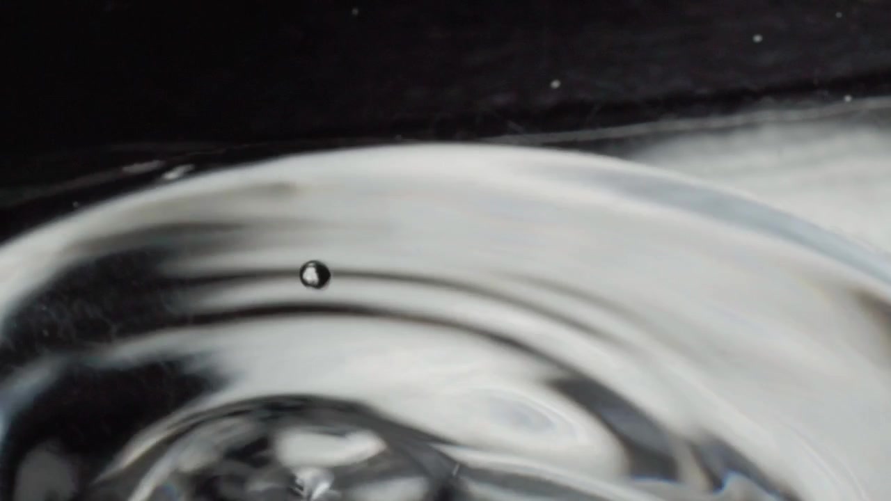 Primo piano macro di una goccia d'acqua che crea un'increspatura circolare sulla superficie liquida al rallentatore. Movimento fluido astratto che forma onde riflettenti con una texture fluida e liscia
