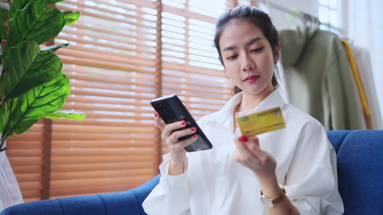 穿着休闲装的亚洲女性，手持智能手机和信用卡进行在线支付，在家享受网上购物的乐趣，面带愉悦笑容。