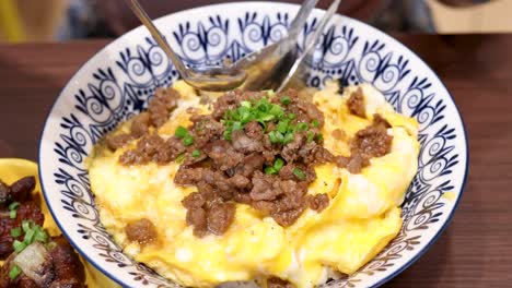 a hand pours fish sauce over a bowl of beef and scrambled egg rice, garnished with herbs, under bright indoor lighting with a close-up perspective