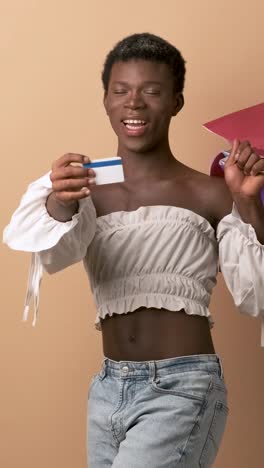 happy transgender person showing a credit card holding shopping bags over beige background