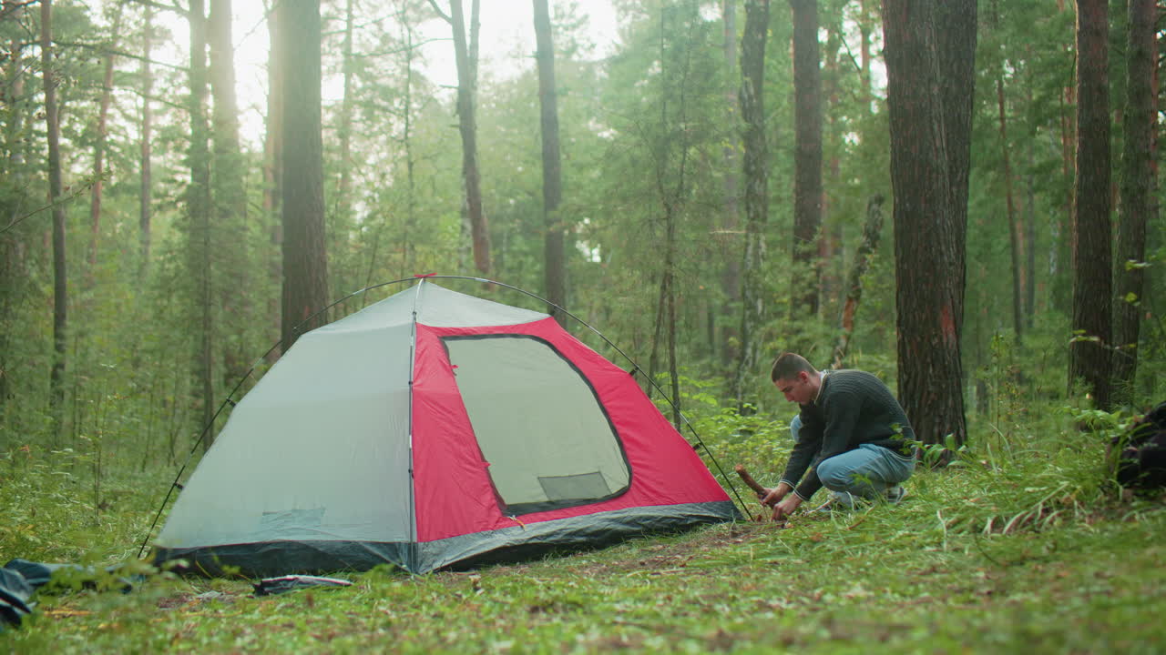 camper squats beside red and gray tent in lush green forest using thick wood to hammer peg into ground during daytime with sunlight filtering through trees and scattered camping gear visible