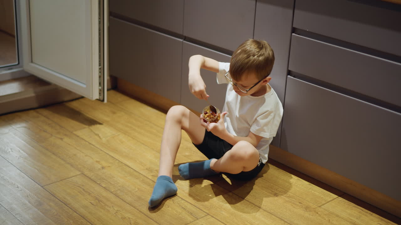 Boy with glasses wearing white shirt and black shorts sitting on wooden kitchen floor near open refrigerator, holding small jar and spoon, eating sweet dessert snack with focus during casual indoor moment