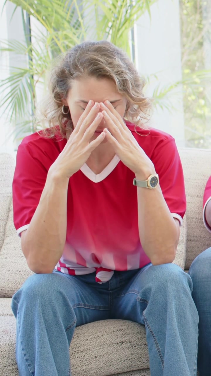 Watching woman in matching sports shirt reacting to offscreen call, covering face, calming on couch