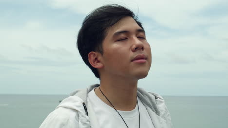 asiatic youg guy relaxing face enjoy summer vibes on the beach with ocean sea in background , slow motion close up portrait