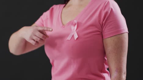 mid section of a woman pointing to the pink ribbon on her chest against black background