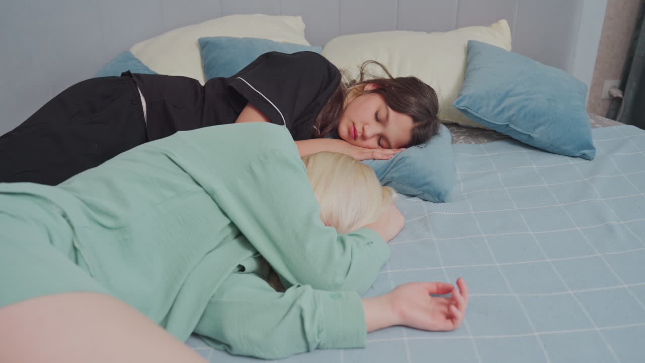 Two women dressed in casual sleepwear rest peacefully on bed covered with soft blue pillows and checkered bedsheet, scene captures calm, restful moment in cozy bedroom setting
