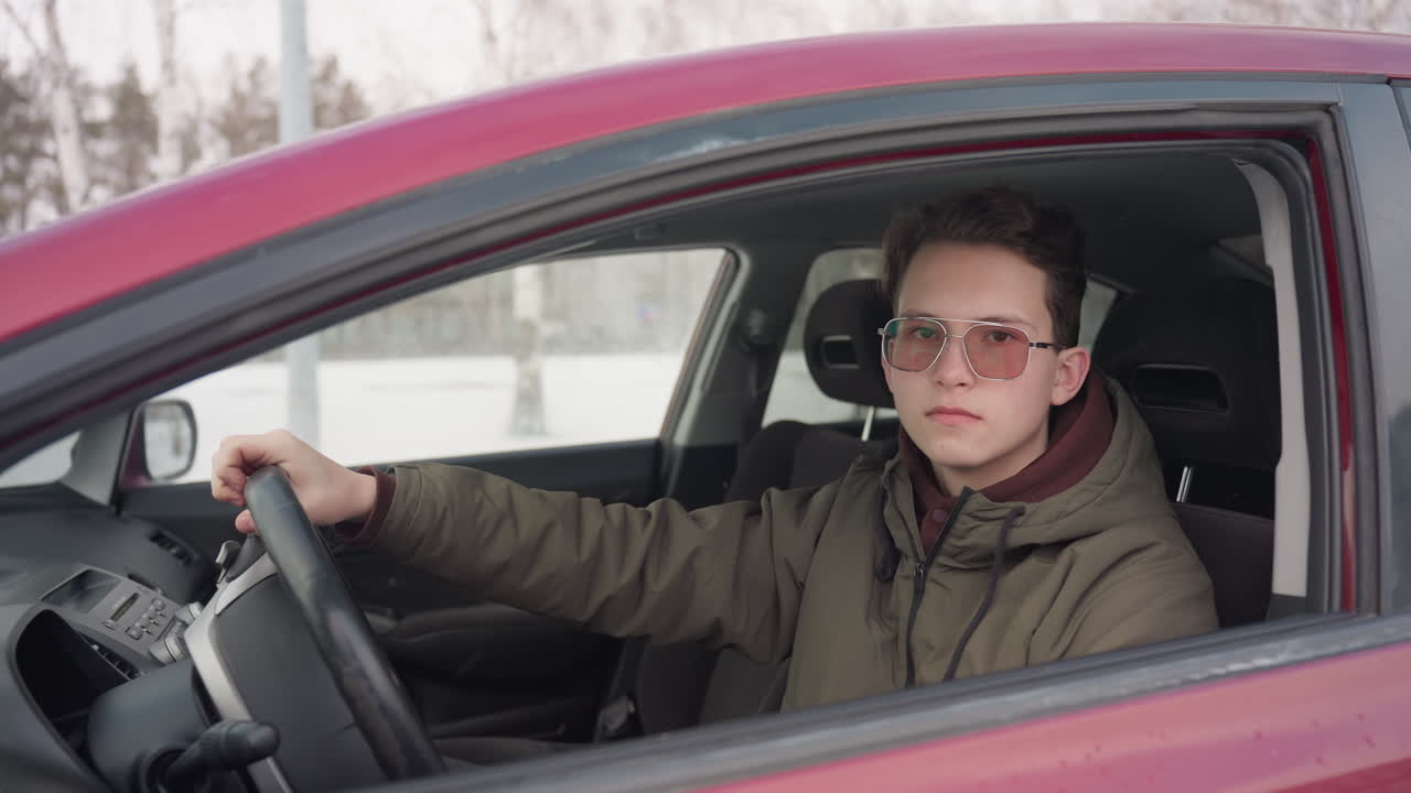 young car owner in winter jacket sitting in red car with hand on steering wheel taps steering while staring outside looking focused surrounded by snow-covered environment on cold winter day