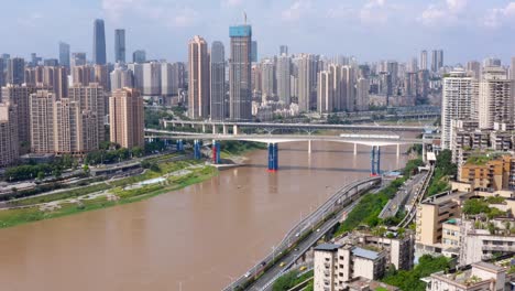 Dolly-push-in-tilt-up-reveals-Chongqing-city-china-skyline-and-murky-brown-river,-blue-sky-day-aerial