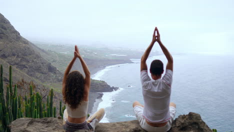 On-a-mountain-peak,-a-man-and-a-woman-meditate-on-stones,-hands-raised,-while-observing-the-ocean-and-engaging-in-relaxation-breathing