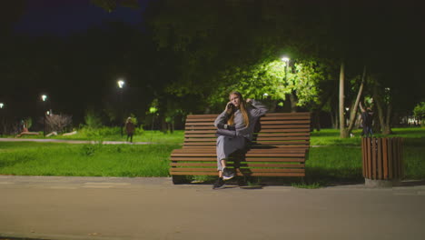 young woman sitting on park bench at night, talking on phone with laptop on lap, resting head on hand, leg crossed, soft lighting with people strolling in background