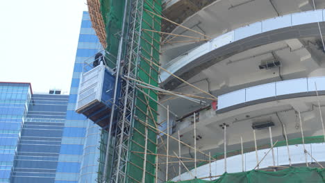 Following-the-Movement-of-Blue-Elevator-and-Cranes-on-a-Hong-Kong-Worksite,-with-Machinery-in-Motion-and-Exterior-Tower-Support