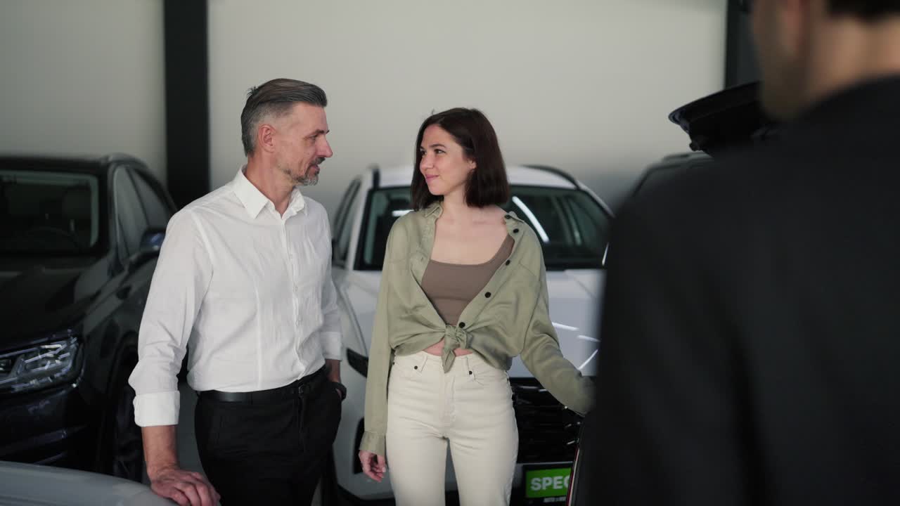 familia confiada en una sala de exhibición de automóviles un hombre de mediana edad con una camisa blanca y su esposa morena se comunican emocionalmente con un asistente en traje de negocios en una salón de exhibiciones de automóviles sobre automóviles modernos