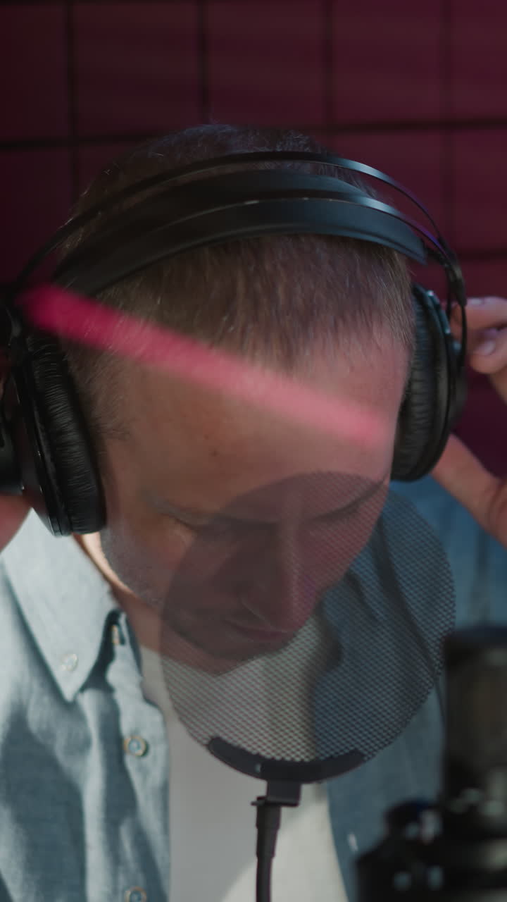 un hombre en un estudio de grabación con una camisa de manga larga azul y auriculares negros, ajustando sus auriculares mientras se prepara para grabar con un fondo de panel acústico rojo, micrófono en primer plano