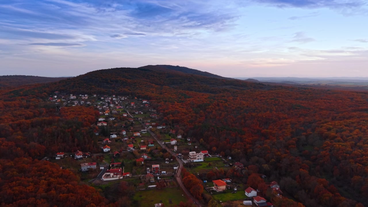 Rural village in an autumn forest valley below a rounded forested hill. At dusk red roofed houses line a valley road enclosed by dense orange autumn forest below a rounded hill.