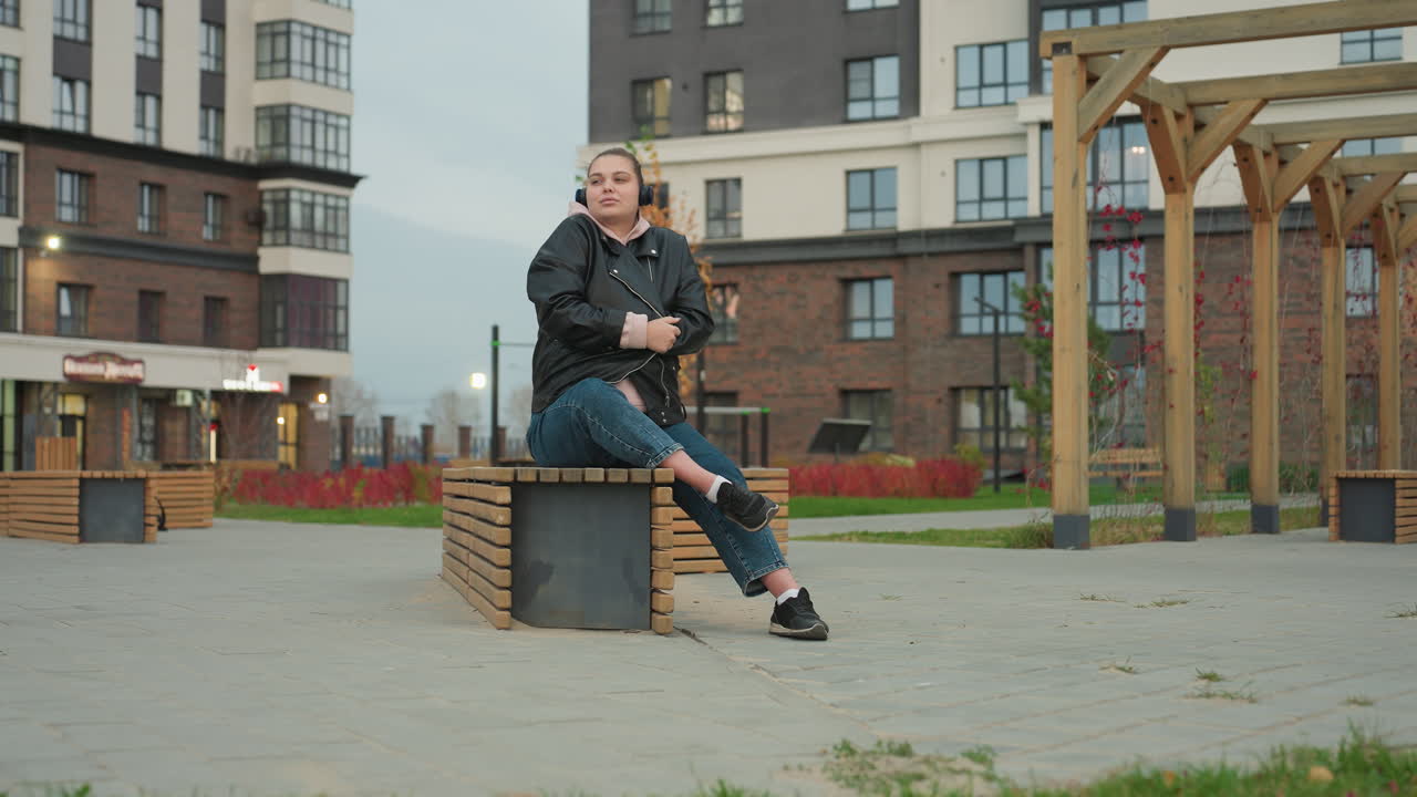 Lady seated on urban wooden bench adjusts jacket to stay warm in cool weather while wearing headphones listening to music outdoors surrounded by modern residential buildings