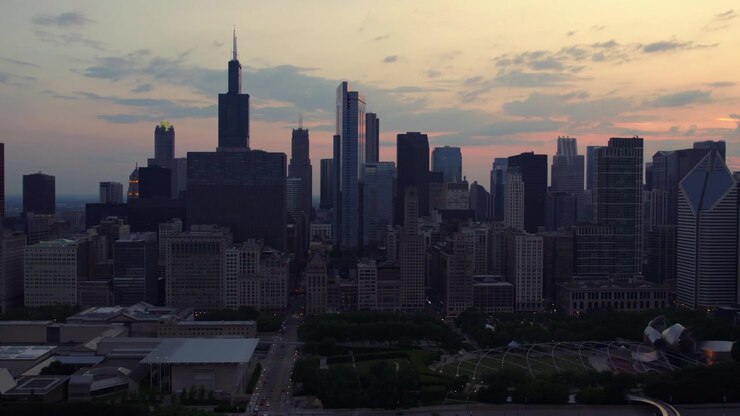Vista aérea ampla de drone do centro de Chicago, apresentando a Willis Tower, arranha-céus densos e um pôr do sol suave em tons de laranja e rosa que se estende pelo céu do entardecer.