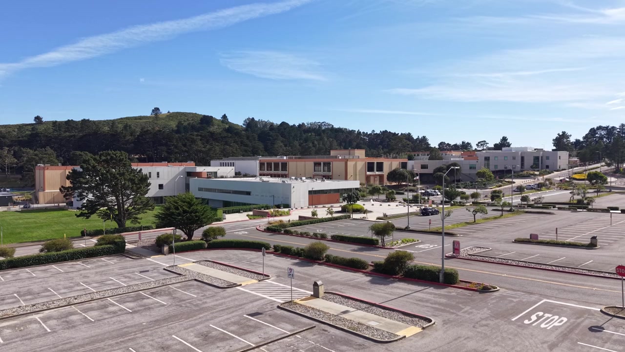 Imágenes de dron aéreas sobrevolando el campus vacío y el estacionamiento de Skyline College en San Bruno, California, mostrando edificios académicos desiertos bajo un cielo azul despejado en un día soleado.