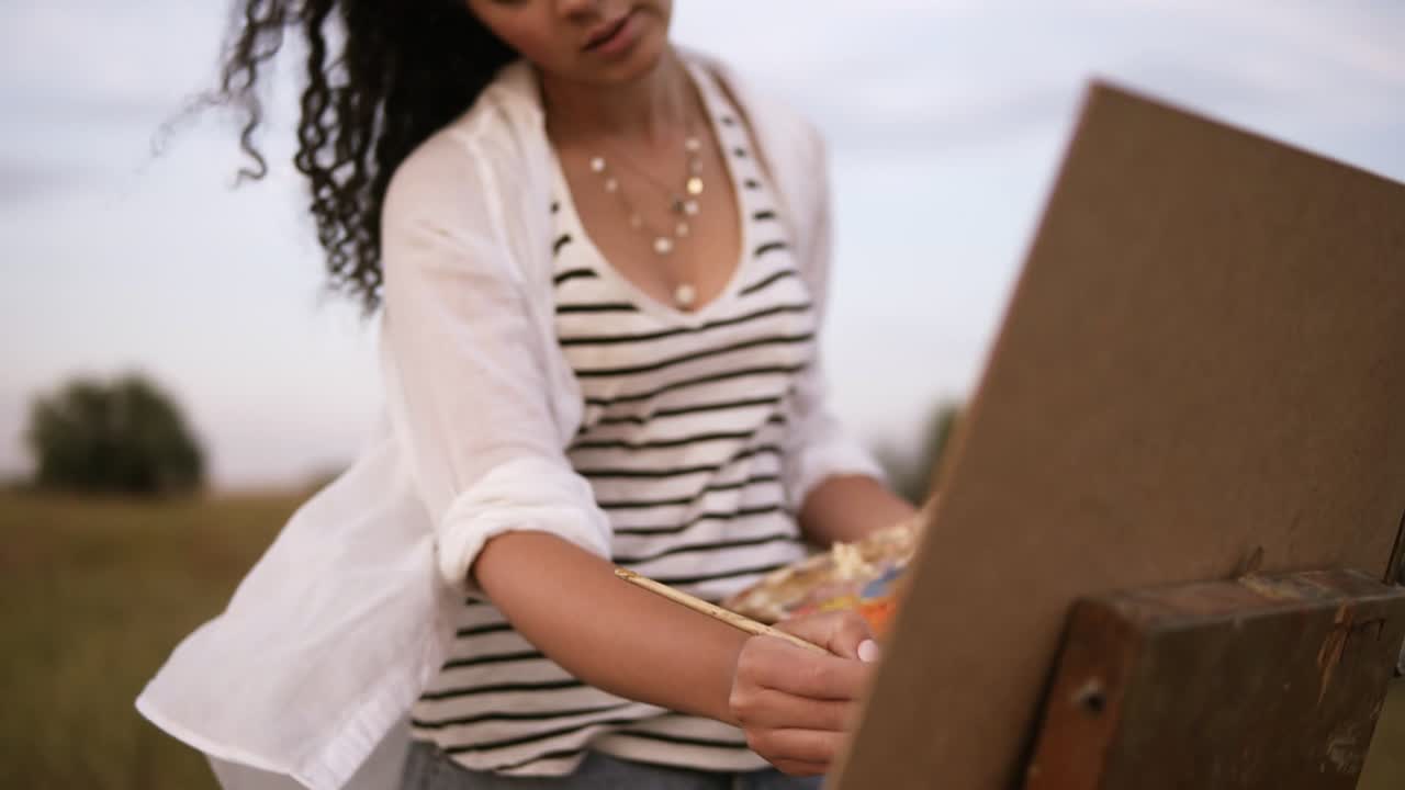 Gorgeous view of a young female artist in work process outdoors. Paints using a palette and easel. Wind waving her curly hair
