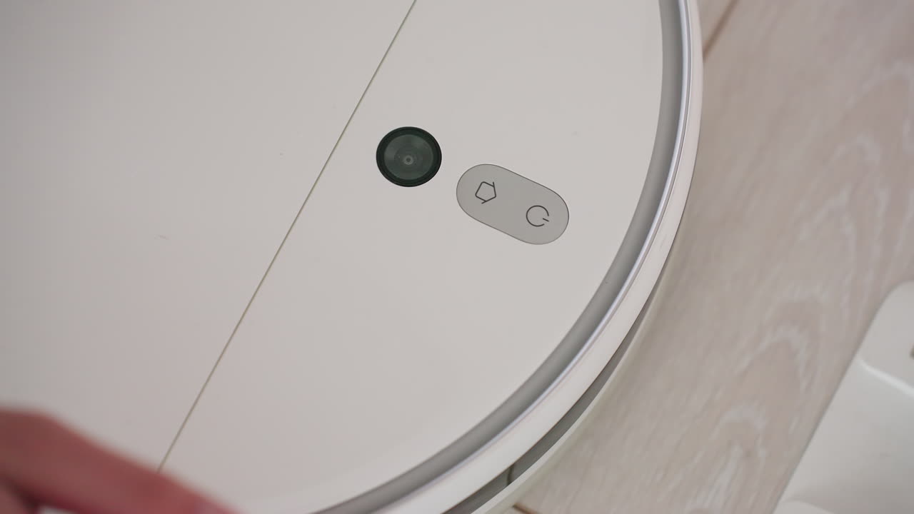 Close up of person switching on robot vacuum, finger pressing control button as device rotates and begins cleaning movement on wooden floor
