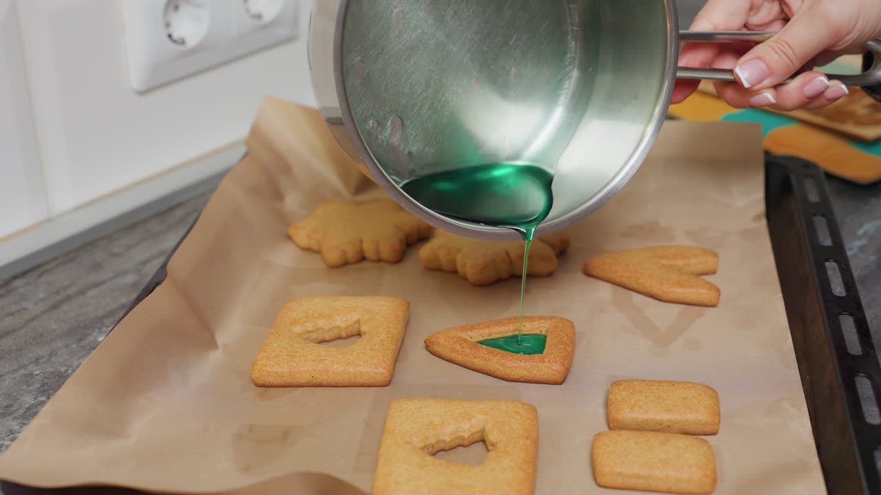 Caterer pouring green liquid from pot over shaped cookies for decoration in modern kitchen, preparing cookies with colorful syrup on parchment paper
