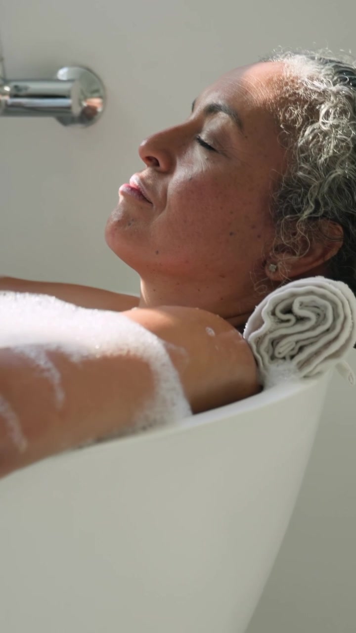 Vertical video: Settling into tub, woman reclining on towel at home, relaxing with foam and faucet