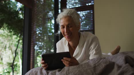 Ältere-Frau-Mit-Gemischter-Abstammung-Liegt-Mit-Tablet-Im-Bett-Und-Lächelt