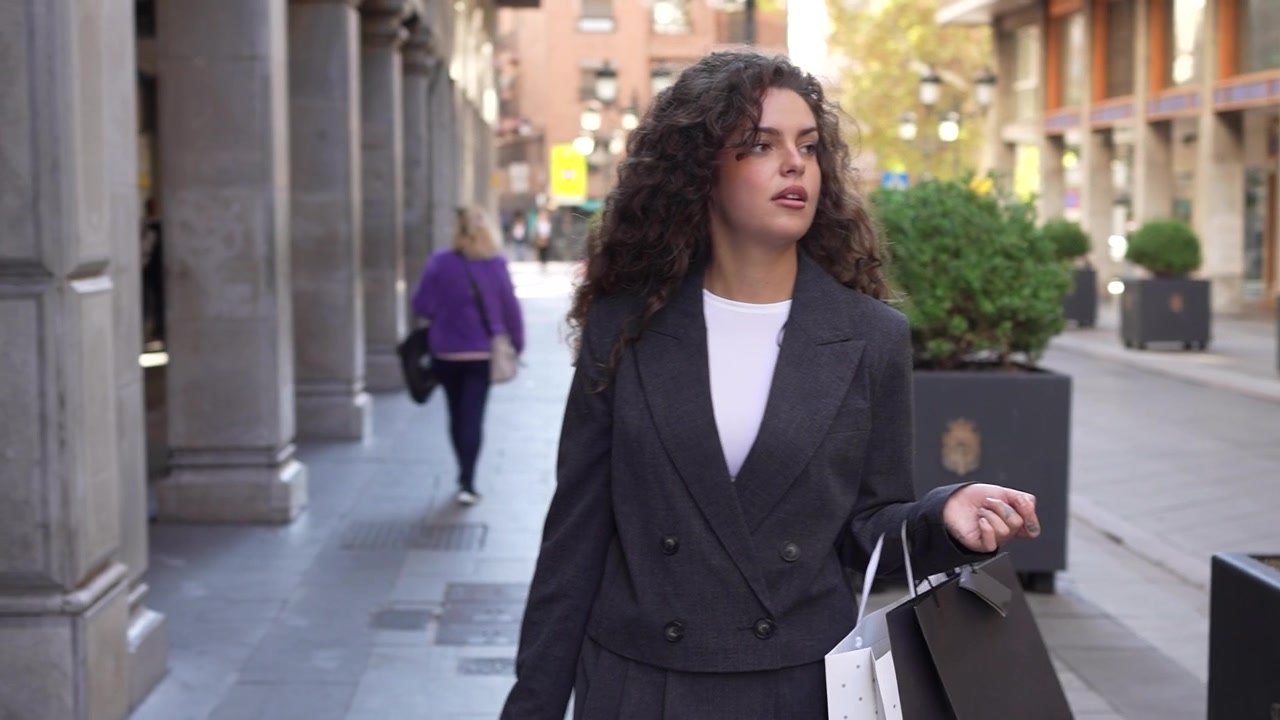Una giovane donna passeggia tranquillamente in un ambiente urbano, con capelli ricci e un outfit alla moda