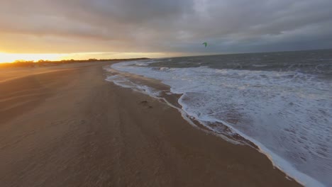 Drone-Siguiendo-La-Playa-Durante-La-Puesta-De-Sol,-Con-Kitesurfistas-En-El-Fondo