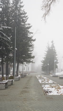 foggy atmosphere with snow-covered light poles, bare trees, and residential buildings lining quiet paved path. fallen leaves, muted tones, and soft lighting create serene wintry
