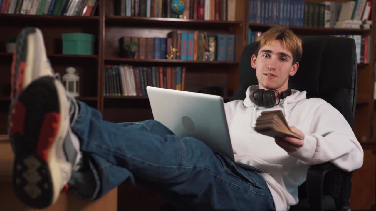 un joven está sentado en una silla con una computadora portátil descansando sobre sus piernas, y está sosteniendo dinero en sus manos