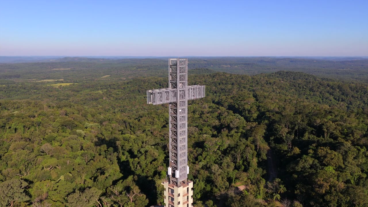 Torre monumental em forma de cruz eleva-se acima do dossel da Mata Atlântica no Parque Temático de la Cruz, Santa Ana, Província de Misiones, Nordeste da Argentina, em imagem aérea de drone com movimento de aproximação durante condições de luz solar.