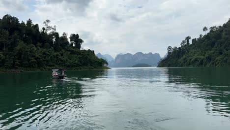 En-El-Lago-Khao-Sok-En-Tailandia,-Un-Barco-Se-Aleja-Suavemente-De-La-Cámara-En-El-Lado-Izquierdo,-Revelando-El-Lago-Y-Las-Montañas-Cubiertas-De-Exuberante-Vegetación-Al-Fondo.