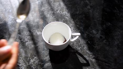 spooning sugar into white cup on black marbled table with shafts of sunlight falling across the scene