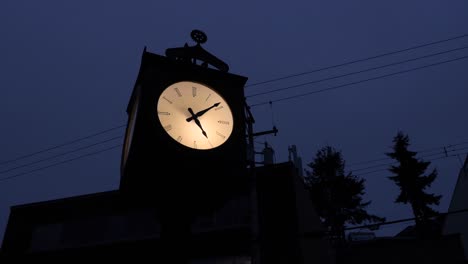 Silhouette-Einer-Alten-Beleuchteten-Standuhr-An-Einem-Dunklen-Abend-Zwischen-Gebäuden-Und-Elektrischen-Leitungen-Am-Himmel