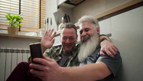 A-happy-brunette-man-with-stubble-and-in-a-green-checkered-shirt-waves-his-hand-Hello-while-sitting-with-his-elderly-boyfriend-with-a-gray-beard-lush-mustache-and-gray-hair-communicating-via-video-conference-with-his-loved-ones-while-sitting-in-the-kitchen-on-the-floor