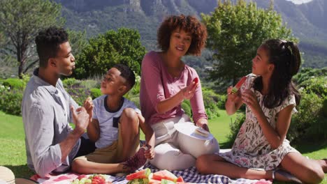 felices padres afroamericanos, hija e hijo sentados al aire libre en una manta, comiendo frutas